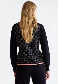 Femme aux longs cheveux blonds portant une veste noire à motifs et un pantalon noir, vue de dos sur fond blanc uni.