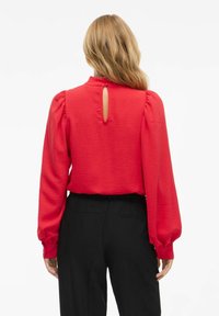 Blusa rossa con maniche lunghe a sbuffo e dettaglio cut-out sulla schiena, realizzata in tessuto testurizzato. Abbinata a pantaloni neri, con minimali dettagli metallici.