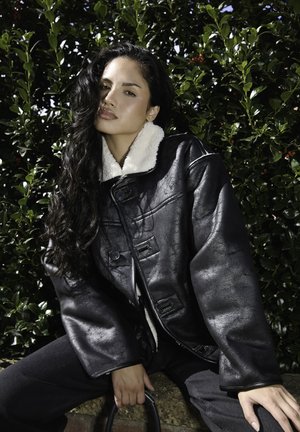 Chaqueta de piel sintética negra con un cuello de borreguito blanco, corte oversize, grandes botones y acabado texturizado. Lleva puesta con vaqueros oscuros, frente a un fondo de vegetación.