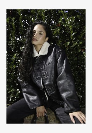 Chaqueta de piel sintética negra con un cuello de borreguito blanco, corte oversize, grandes botones y acabado texturizado. Lleva puesta con vaqueros oscuros, frente a un fondo de vegetación.