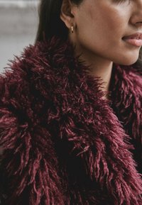 Bordeauxfarbener Kunstfellschal mit langem, zotteligem Stil. Nahaufnahme zeigt Detail und Weichheit, ergänzt durch einen kleinen goldenen Creolen-Ohrring.