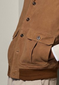 Braune Wildlederjacke mit Knopfleiste, zwei Taschen mit Klappen und geripptem Saum. Glatte Textur und taillierte Passform sind sichtbar.