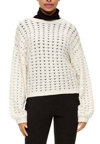 Weißer Strickpullover mit lockerem, offenem Strickmuster, langen Ärmeln und geripptem Saum. Über einem schwarzen Rollkragenpullover und schwarzen gerippten Hosen getragen.