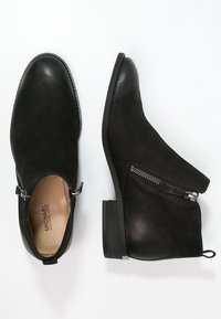 Bottines en suède noir avec un bout rond, fermeture éclair sur le côté et petit talon carré. Dotées de coutures discrètes et d'une boucle décorative sur le côté.