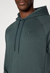 Tmavě zelená mikina s kapucí vyrobená z měkké textilie, která má přední klokaní kapsu a šňůrku na kapuci. Obsahuje malé černé logo Nike na hrudi.
