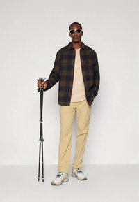 Geruite flanellen blouse in donker groen en marineblauw, beige T-shirt, lichtbruine broek en grijze sneakers. Houdt een zwarte wandelstok vast.