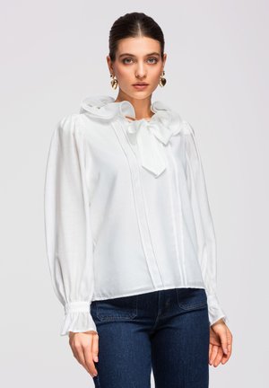 Femme portant un chemisier blanc à col à volants et nœud, associé à un jean bleu foncé, sur un fond clair uni.