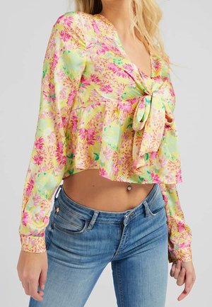 Kobieta w żółtej kwiatowej bluzce z długim rękawem i krótkim krojem z wiązaniem z przodu oraz niebieskich dżinsach na jednolitym tle.