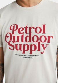 Creme bomulds T-shirt med fed rød tekst, der siger "Petrol Outdoor Supply", med yderligere detaljer i mindre blå tekst nedenfor.