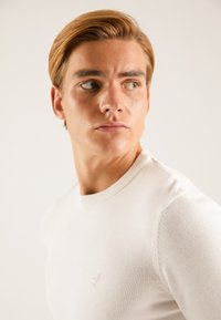 Pull blanc à col rond avec un col côtelé. Tissu en maille léger, avec un logo subtil sur la poitrine, texture lisse, coupe ajustée.