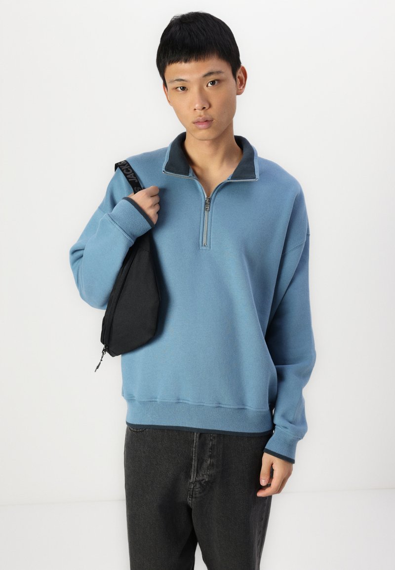 Lichtblauwe sweatshirt met een halve rits kraag en geribde manchetten. Model houdt een zwarte schoudertas vast. Eenvoudig ontwerp, zachte textuur.