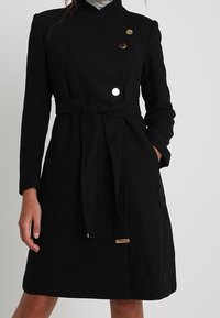 Abrigo de lana negro con diseño envolvente, tres botones en contraste, un cinturón atado en la cintura y bolsillos laterales. Textura suave, hasta la rodilla.