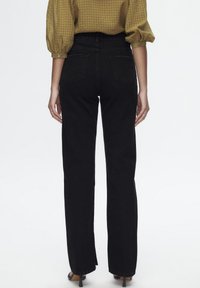 Jeans noirs taille haute avec une coupe droite, dotés de poches arrières et de détails de couture subtils. Le mannequin porte une blouse à carreaux.