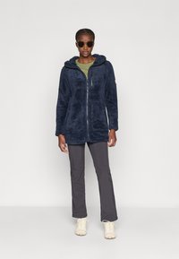Giacca in pile di colore blu scuro con cappuccio, zip frontale e due tasche laterali. Abbinata a pantaloni grigio scuro e scarpe beige chiare.