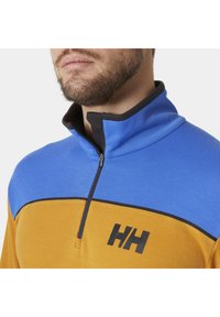 Półzapinany sweter w pomarańczowym i niebieskim kolorze, z czarnym zamkiem błyskawicznym i logo. Miękka tkanina, usztywniony kołnierz, z designem w blokach kolorystycznych.