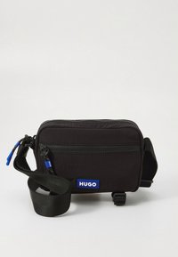 Small black crossbody bag with adjustable strap, front zip pocket, and blue "HUGO" logo patch on plain background.
