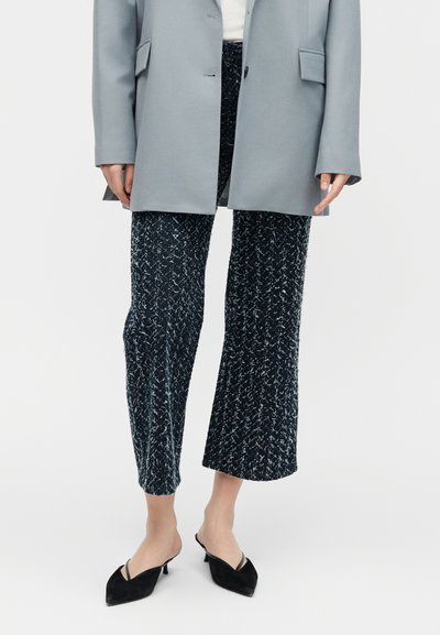 Femme portant un pantalon large court noir et blanc tacheté, un blazer gris et des mules noires à talons kitten pointus.