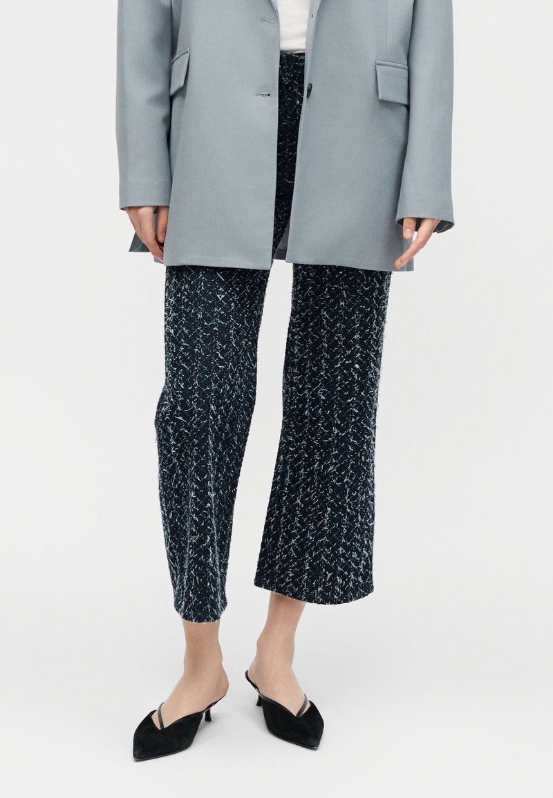 Femme portant un pantalon large court noir et blanc tacheté, un blazer gris et des mules noires à talons kitten pointus.