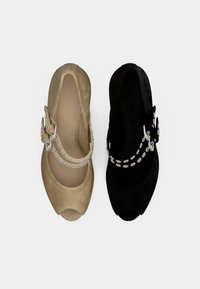 Δύο ζευγάρια παπούτσια με ανοιχτά δάχτυλα: ένα μπεζ σουέντ, ένα μαύρο σουέντ. Κάθε ένα έχει ένα διακοσμητικό λουράκι με κυκλικές ασημένιες λεπτομέρειες.