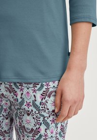 Blauwe, zachte stoftop met driekwart mouwen; gecombineerd met lichtblauwe broek met een patroon van bloemen en bladeren in paars en bordeaux.