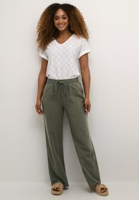 White V-neck top with eyelet pattern, paired with olive green linen drawstring pants, and light tan braided sandals.