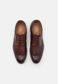 Chaussures habillées en cuir marron avec des détails en brogue, présentant une finition lisse, un bout rond et un design à lacets. Nom de la marque "Clarks" visible à l'intérieur.
