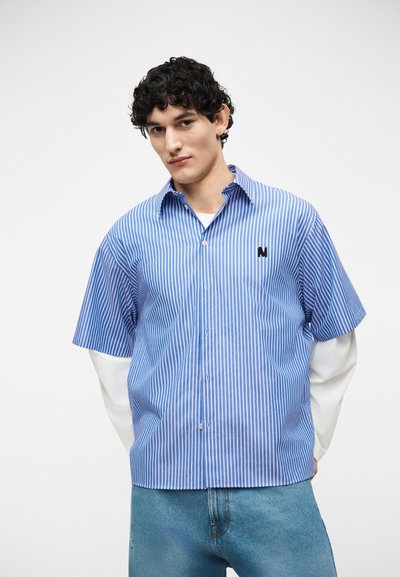 Jeune homme aux cheveux bouclés portant une chemise à manches courtes rayée bleue et blanche superposée sur une chemise blanche à manches longues et un jean bleu.