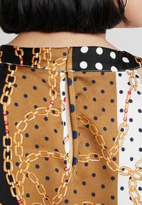 Blusa marrone a fantasia con grafiche di catene dorate e pois neri. Include una piccola fessura sul retro e un design del colletto a contrasto.