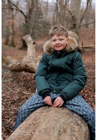 Green, padded jacket with a fur-lined hood, paired with blue pants featuring a leaf pattern. Sitting on a log in a forest setting.