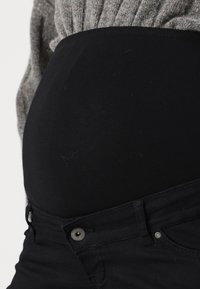 Primer plano de una barriga embarazada vistiendo unos vaqueros de maternidad negros y un suéter gris.