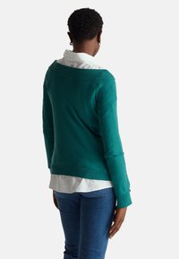 Tyrkysový pletený svetr s širokým výstřihem a žebrovanými manžetami, navrstvený přes bílou košili s límečkem, nošený s modrými džíny.