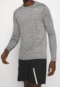 Langärmliges, graues Sportshirt mit strukturiertem Finish und einem weißen Logo auf der Brust, kombiniert mit schwarzen Shorts mit einem gelben Akzentstreifen.
