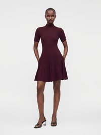 Femme debout les mains dans les poches portant une robe côtelée rouge foncé à manches courtes et des talons ouverts noirs sur un fond uni.