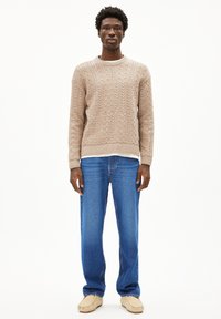 Pull en maille beige à manches longues, col rond et poignets côtelés, associé à un jean bleu droit et des chaussures beiges à enfiler.