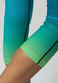 Dopasowane legginsy sportowe sięgające do kostek w gradientzie od turkusu do zieleni. Materiał jest gładki, z czarnymi szwami wzdłuż wszystkich krawędzi.