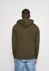 Sudadera verde oliva hecha de una suave mezcla de algodón, con capucha ajustable, puños acanalados y un corte holgado, combinada con jeans de color azul claro.