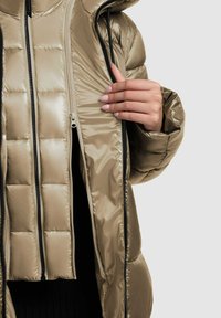 Jachetă căptușită de culoare bej metalic, cu un finisaj lucios. Dispune de închidere cu fermoar, glugă și un model matlasat vizibil.