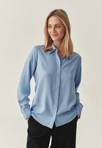 Cămașă lungă, de culoare albastră deschisă, cu guler cu butoni, textură netedă și tiv curbat. Asortată cu pantaloni negri.