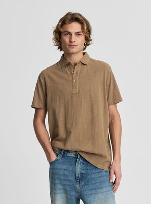 Giovane uomo con capelli biondi ondulati indossa una polo marrone a maniche corte e jeans blu, in piedi con una mano in tasca.