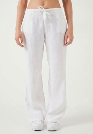 Kobieta ubrana w luźne, białe, teksturowane spodnie z sznurkiem w pasie i białe trampki, stoi na tle jednolitego tła.