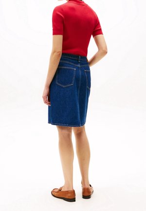 Vrouw draagt een rood kortärmelig gebreid shirt, een blauwe spijkerrok tot de knie en bruine loafers, staat met haar rug naar de camera gericht.