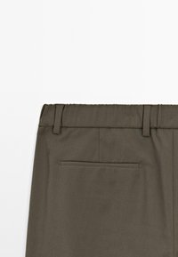 Mörk olivgrön byxlinning med bältesslingor och en enkel passpoal bakficka på vit bakgrund.
