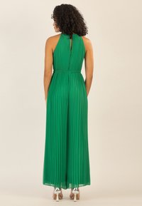 Vestido verde plisado de tirantes con espalda de llave y cinturilla elástica, con una silueta fluida y combinado con zapatos de tacón alto plateados.