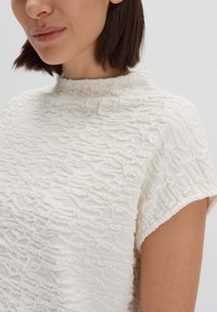 Blusa bianca, con texture, colletto alla coreana e maniche corte, caratterizzata da un motivo ondulato sulla stoffa.