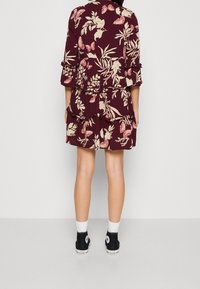 Burgundy blommig klänning med volanger, prydd med rosa och krämfärgad botanisk mönster; i kombination med svarta sneakers med hög skärning och vita strumpor.