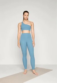 Jovem mulher em pé descalça sobre um tapete de yoga, vestindo um top desportivo azul de um ombro só e leggings de cintura alta, contra um fundo simples.