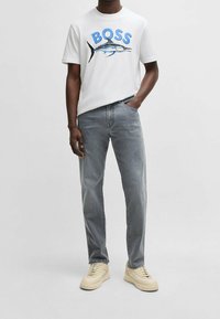 Vit t-shirt med en blå "BOSS" logga och en grafisk fiskdesign, tillsammans med ljusgrå denimjeans och krämfärgade sneakers.