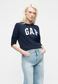 Νεαρή γυναίκα με ξανθά μαλλιά, φορώντας ένα μπλε σκούρο φούτερ GAP και ανοιχτόχρωμο μπλε τζιν, με τα χέρια στις τσέπες, στέκεται μπροστά σε μια απλή φόντο.