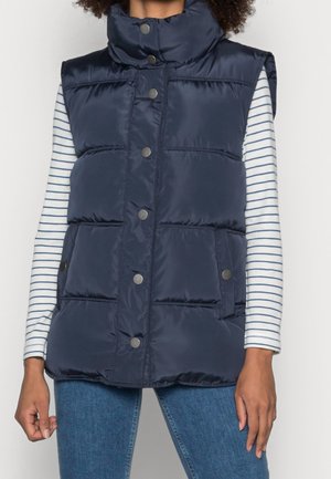 Nainen, joka pukeutunut laivastonsiniseen tikattuun hiuksettomaan toppaliiviin, jossa on painonapit, valkoisen ja siniraidallisen pitkähihnaisen paidan sekä farkkujen päällä.