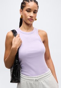 Νεαρή γυναίκα με πλεγμένα μαλλιά, φορώντας χρυσούς κρίκους στα αυτιά, λιλά αμάνικη μπλούζα, γκρι φόρμες και κρατώντας μαύρη τσάντα στον ώμο.